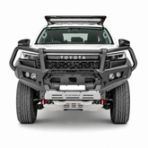 A front-facing studio shot of a white Toyota Hilux pickup truck bakkie equipped with a heavy-duty black Rockarmor bull bar, silver front bumper, roof-mounted LED light bar, and off-road tires on a light gray background.