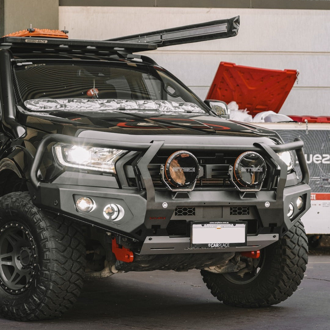A black off-road truck with a heavy-duty black front bumper guard, dual round auxiliary lights, and a roof-mounted light bar, photographed in an outdoor or semi-outdoor setting.