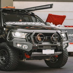 A black off-road truck with a heavy-duty black front bumper guard, dual round auxiliary lights, and a roof-mounted light bar, photographed in an outdoor or semi-outdoor setting.