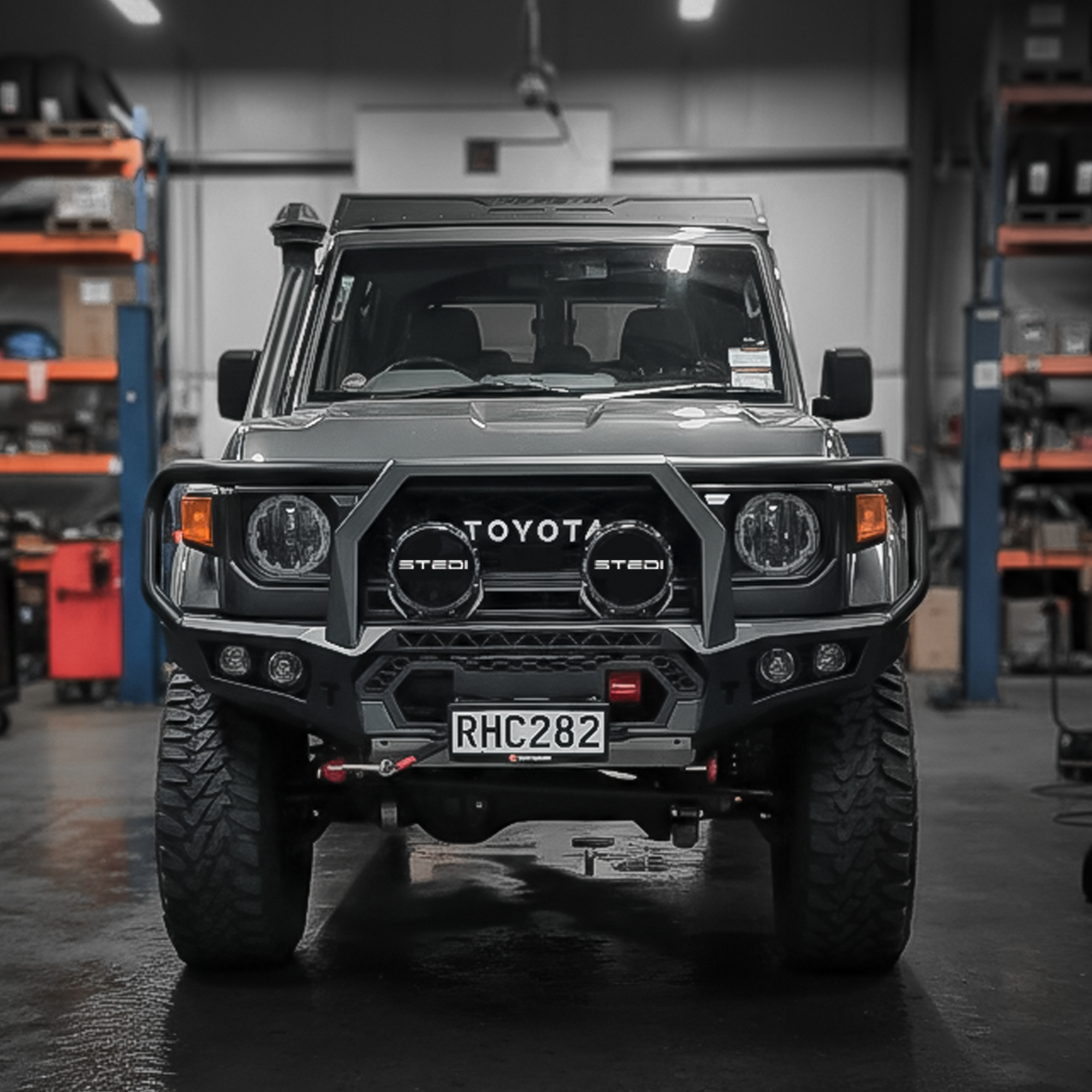 A black Toyota Land Cruiser with a heavy-duty front bull bar, dual round auxiliary lights, and a custom license plate, parked in a workshop with shelves in the background.