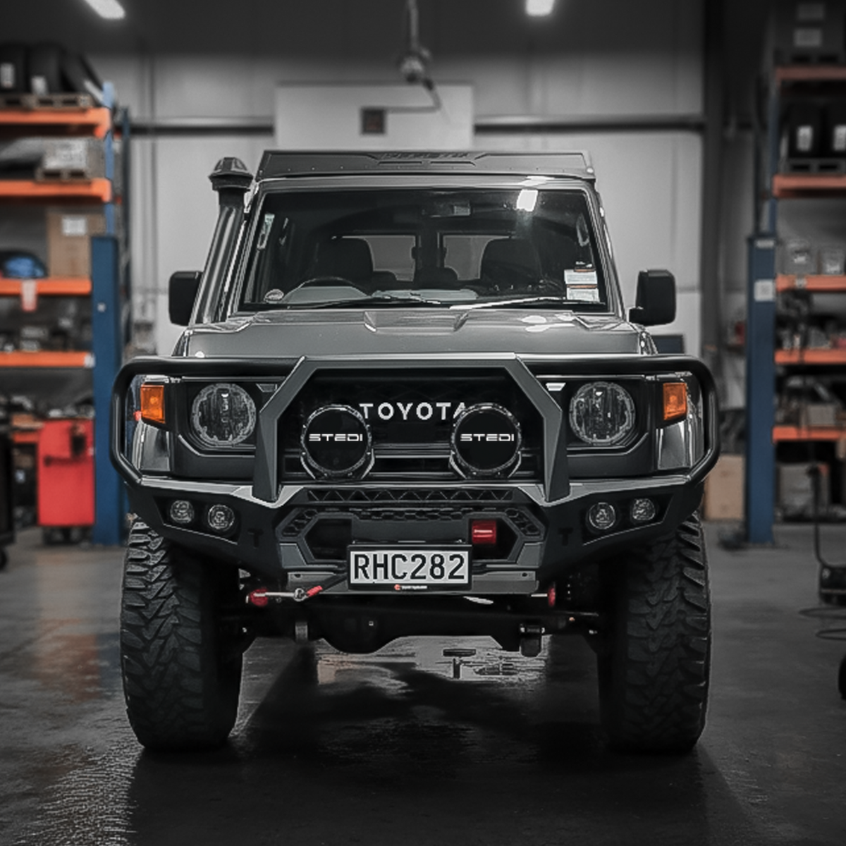 A black Toyota Land Cruiser with a heavy-duty front bull bar, dual round auxiliary lights, and a custom license plate, parked in a workshop with shelves in the background.