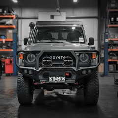 A black Toyota Land Cruiser with a heavy-duty front bull bar, dual round auxiliary lights, and a custom license plate, parked in a workshop with shelves in the background.
