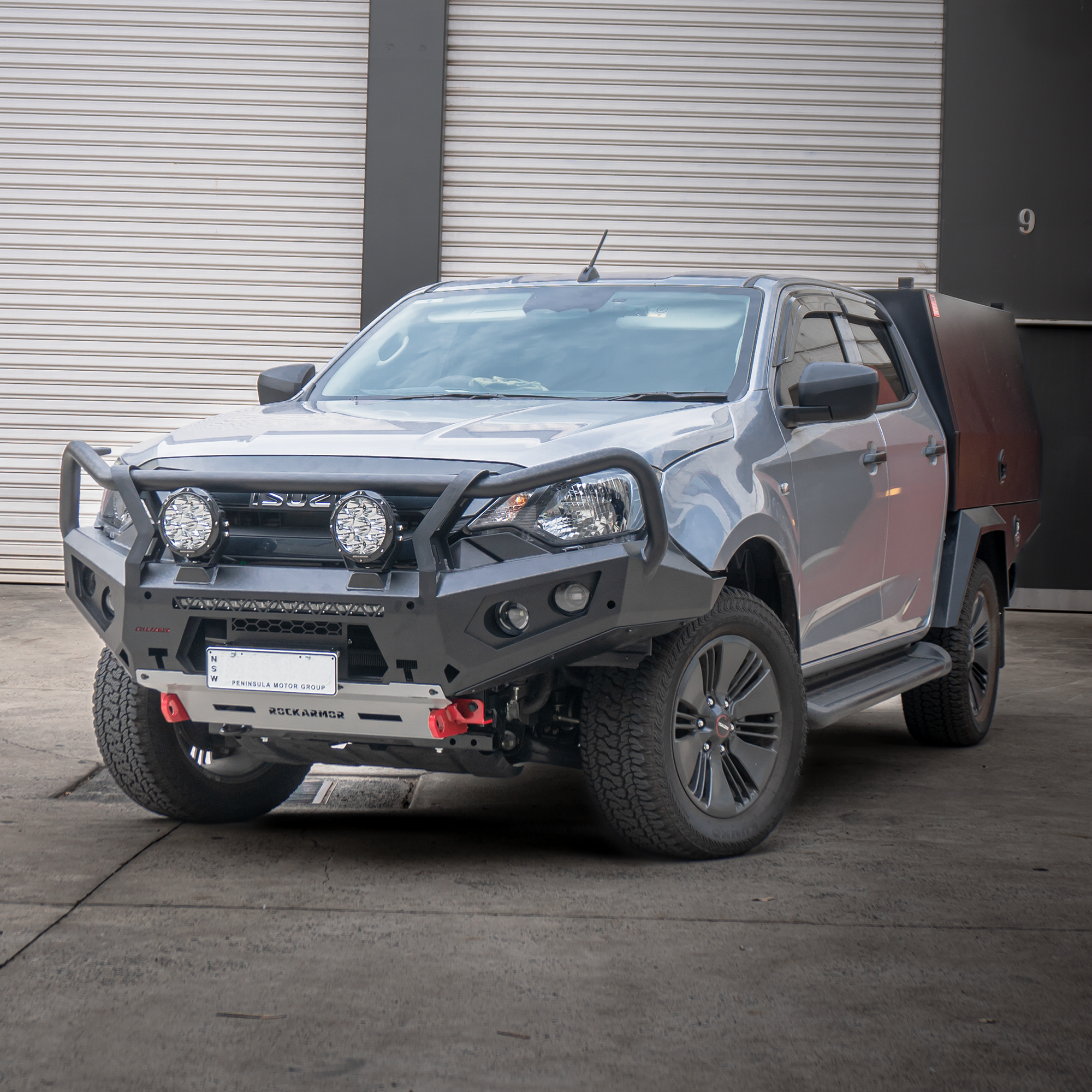 Silver bakkie with additional front bumper bull bar and lights in front of a gray building.