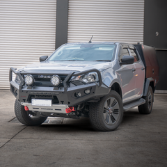 Silver bakkie with additional front bumper bull bar and lights in front of a gray building.