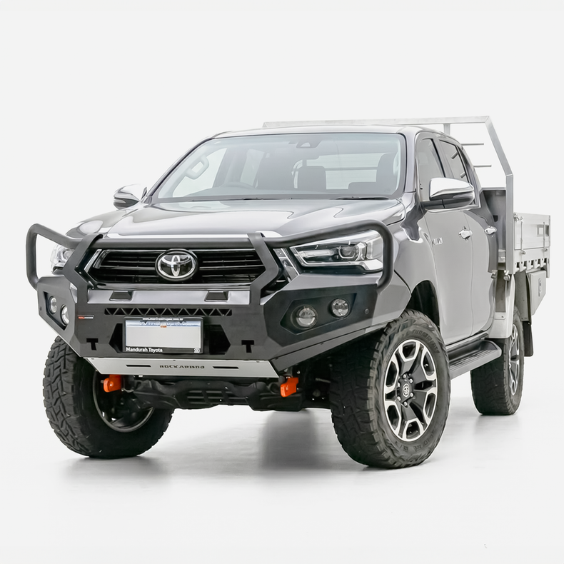 A dark gray Toyota Hilux bakkie pickup truck fitted with a heavy-duty Rockarmor black front bull bar and a silver metal flatbed carrier, photographed on a seamless white studio background.