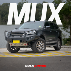A black Isuzu SUV with a heavy-duty front bumper and bull bar, parked outdoors near water with large white 'M-UX' text overlay and the 'ROCKARMOR' brand logo at the bottom.