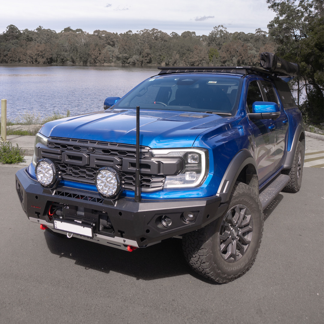 A blue Ford Ranger pickup truck bakkie with a black front bumper guard, roof rack, and off-road tires, parked outdoors next to a lake with trees in the background.