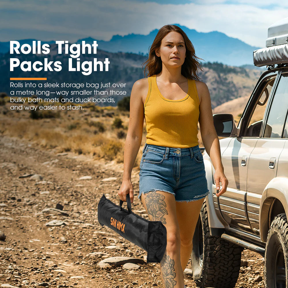 Woman walking in a desert landscape with a vehicle, holding a black SAN HIMA carry bag for a shower tent base awning floor.