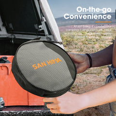 Person holding a gray travel case for a camping shower with 'SAN HIMA' branding, outdoors. 