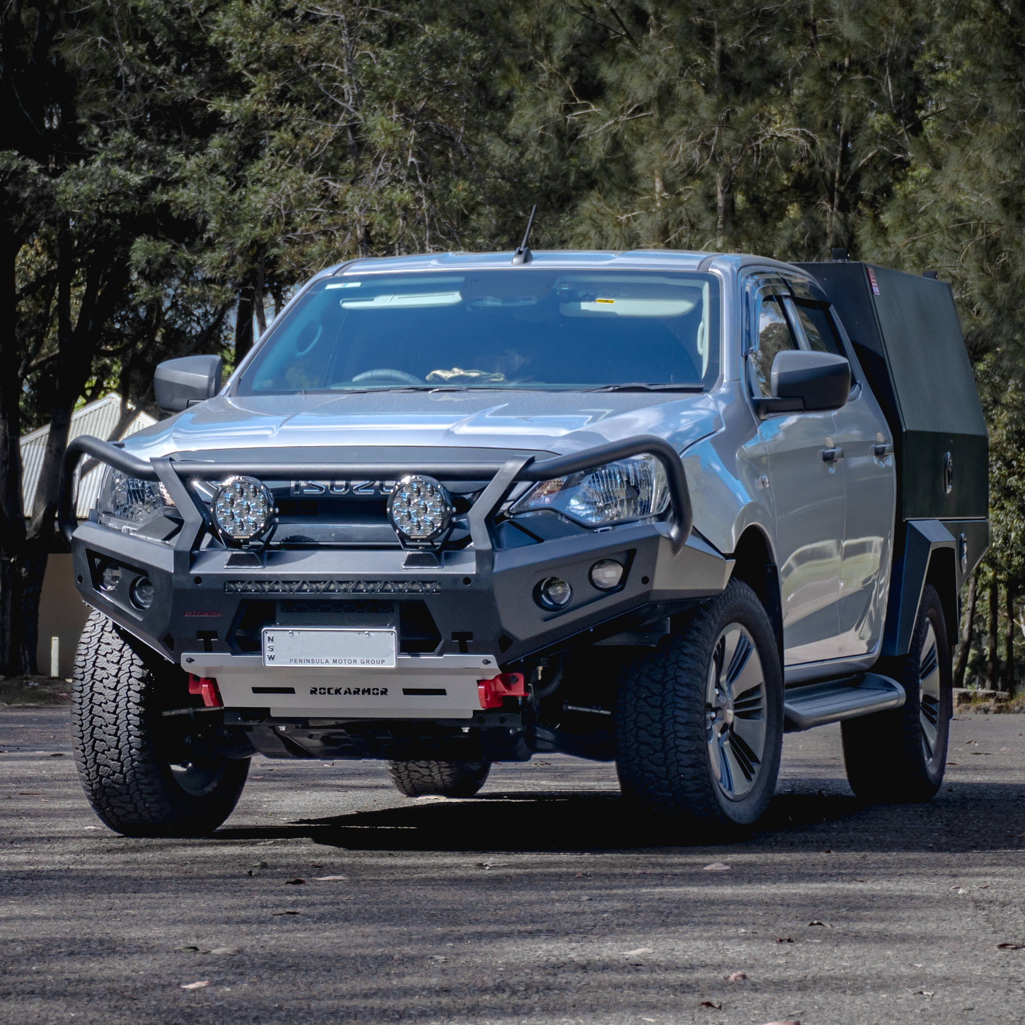 Silver Isuzu bakkie pickup truck with a bull bar and camper shell on a road with trees in the background