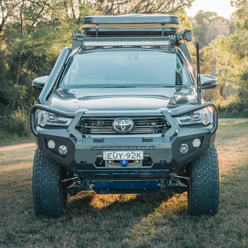 A front-facing view of a dark gray Toyota Hilux bakkie pickup truck parked outdoors on grass with trees in the background. The truck is heavily modified with a large black front bumper bull bar manufactured by Rockarmor, off-road tires, a roof rack with a cargo box, and a light bar.