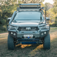 A front-facing view of a dark gray Toyota Hilux bakkie pickup truck parked outdoors on grass with trees in the background. The truck is heavily modified with a large black front bumper bull bar manufactured by Rockarmor, off-road tires, a roof rack with a cargo box, and a light bar.