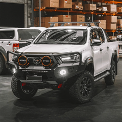 White Toyota Hilux bakkie pickup truck Rockarmor bull bar in a warehouse setting with other vehicles and stacked boxes in the background.