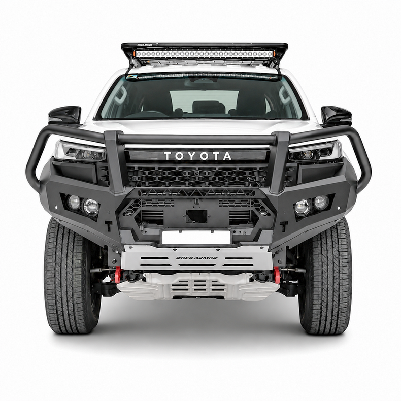 A front-facing studio shot of a white Toyota Hilux pickup truck bakkie equipped with a heavy-duty black Rockarmor bull bar, silver front bumper, roof-mounted LED light bar, and off-road tires on a light gray background.