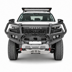 A front-facing studio shot of a white Toyota Hilux pickup truck bakkie equipped with a heavy-duty black Rockarmor bull bar, silver front bumper, roof-mounted LED light bar, and off-road tires on a light gray background.
