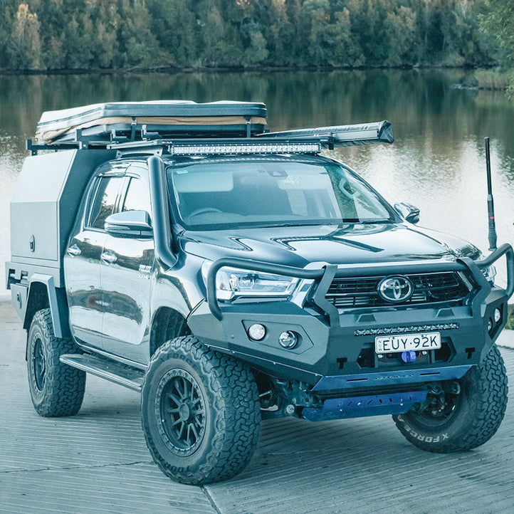 A dark gray Toyota Hilux pickup truck bakkie with a custom off-road cab cover, roof rack, and Rockarmor front bumper bull bar guard parked on a wooden dock beside a lake.