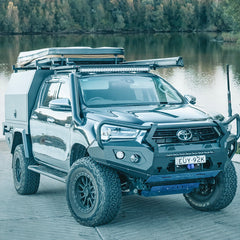 A dark gray Toyota Hilux pickup truck bakkie with a custom off-road cab cover, roof rack, and Rockarmor front bumper bull bar guard parked on a wooden dock beside a lake.