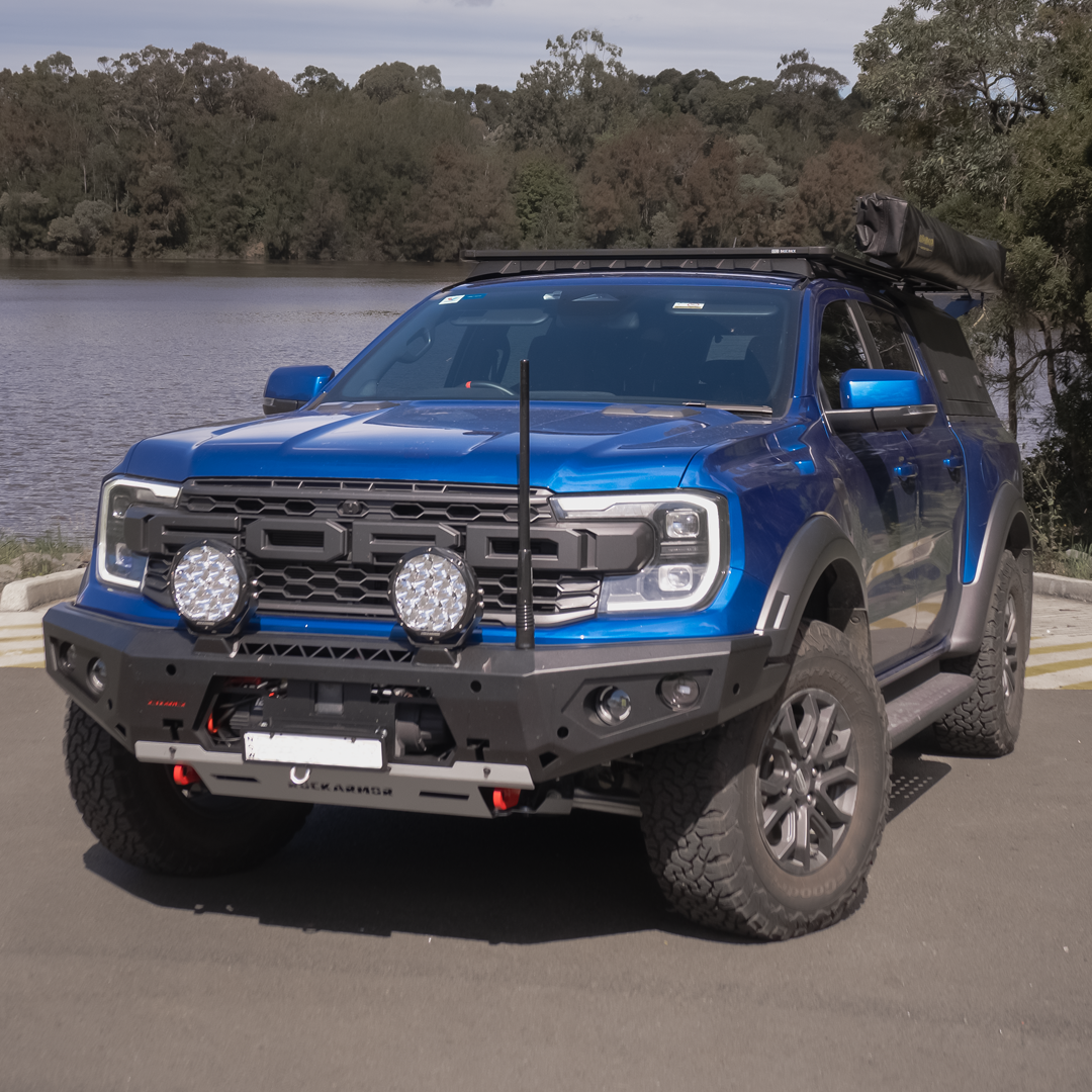 A blue Ford Ranger pickup truck bakkie parked outdoors near a lake, shown from the front three-quarter angle. The truck is equipped with a black off-road front bumper, dual round auxiliary lights, a roof rack with a rolled tent, and off-road tires.