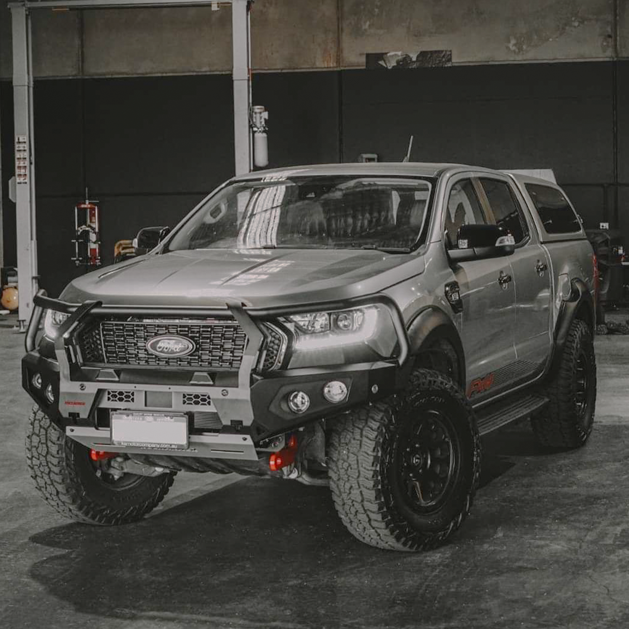 A Ford Ranger pickup truck bakkie with a Rockarmor black front bumper bull bar, off-road tires, and a gray cap, photographed in a garage setting.