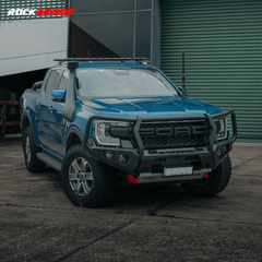 A blue Ford Ranger pickup truck bakkie parked outdoors in front of a green corrugated metal garage door, fitted with a heavy-duty Rockarmor black front bumper bull bar guard and roof rack.