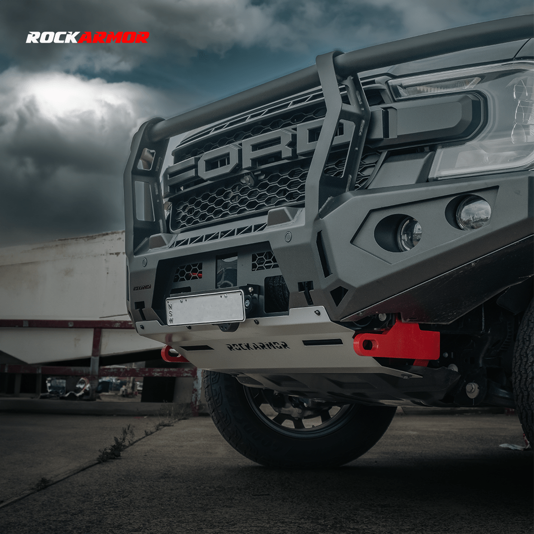 A close-up of a dark gray Ford off-road truck with a heavy-duty front bumper and bull bar by Rockarmor, featuring red tow hooks, set against a cloudy sky and industrial background.
