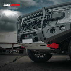 A close-up of a dark gray Ford off-road truck with a heavy-duty front bumper and bull bar by Rockarmor, featuring red tow hooks, set against a cloudy sky and industrial background.