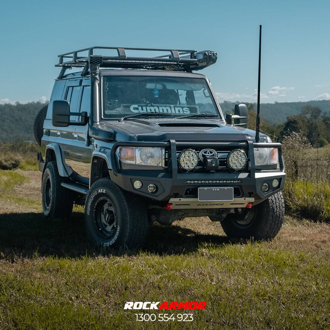 A black Toyota Land Cruiser SUV with a roof rack, front bumper guard, and auxiliary lights, parked outdoors on a grassy field under a blue sky. A decal is visible on the windshield, and a 'Rockarmor' logo watermark is at the bottom.