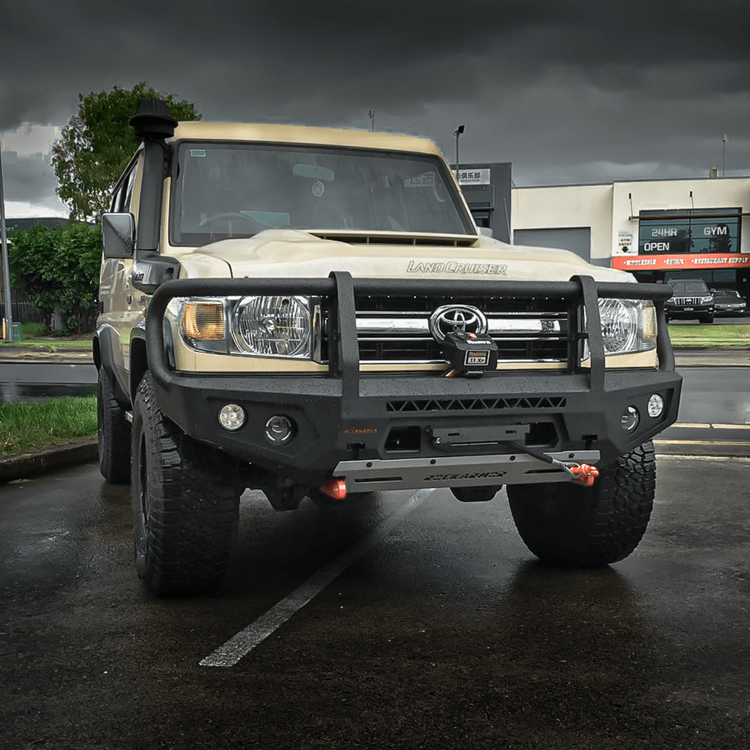 A lifted beige Toyota Land Cruiser with a heavy-duty black bull bar and off-road tires, parked on a wet asphalt lot under a dark, cloudy sky.