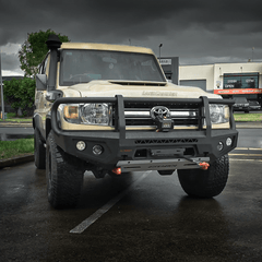 A lifted beige Toyota Land Cruiser with a heavy-duty black bull bar and off-road tires, parked on a wet asphalt lot under a dark, cloudy sky.