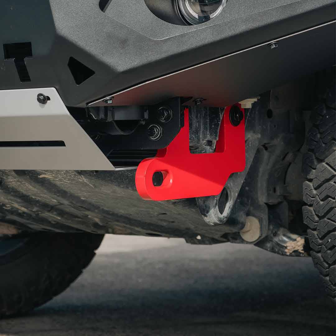 A bright red metal bull bar bracket mounted on the underbody of a dark gray vehicle, shown in a close-up outdoor shot with tires and ground visible.