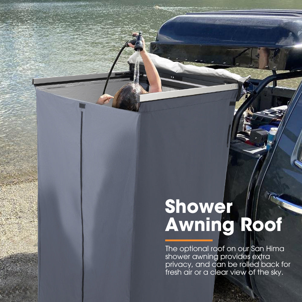 Person using a San Hima portable shower with an awning roof next to a vehicle by a body of water.