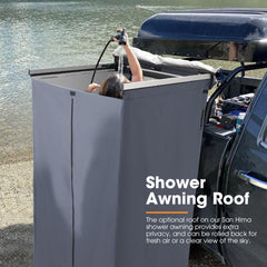 Person using a San Hima portable shower with an awning roof next to a vehicle by a body of water.